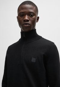 Zwarte zip-up trui van gebreid materiaal, met een hoge kraag en een klein zwart vierkant logo op de borst. Gladde textuur, slim fit.