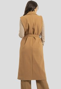 Un manteau sans manches marron avec une ceinture, fait d'un tissu texturé. Il présente un col cranté et une silhouette fluide, porté sur un top gris.