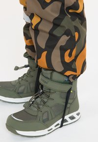Grønne vandtætte støvler med mesh-detaljer og sorte snørebånd. Parret med bukser i orange og camouflage-mønster. Hvide såler som accent.