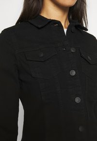 Svart denimjacka med knäppning fram, dubbla bröstfickor och krage. Materialet har en slät textur med synliga sömdetaljer.