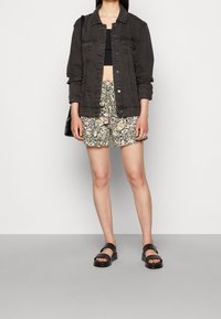 Chaqueta de mezclilla negra con cierre de botón, pantalones cortos estampados de flores en rosa y verde, y sandalias negras. La modelo sostiene un bolso negro de hombro.