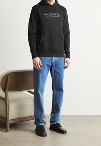 Felpa nera con cappuccio e logo, tasca frontale, abbinata a jeans blu a gamba dritta e scarpe nere. Il modello è in piedi vicino a una sedia di legno.