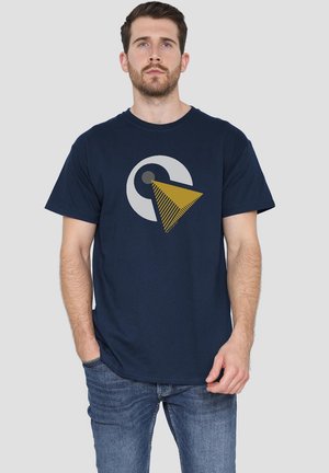 Mann mit kurzen braunen Haaren und Bart, der ein navyblaues T-Shirt mit einem grauen Kreis und goldenen dreieckigen Design trägt, kombiniert mit blauen Jeans, steht vor einem grauen Hintergrund.