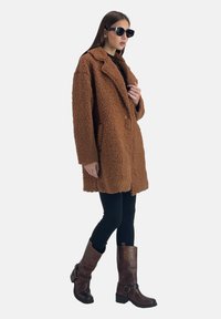 Bruine pluizige jas met een reverskraag, zwarte legging en kniehoge bruine laarzen met gespen. Zwarte vierkante zonnebril maakt de look compleet.