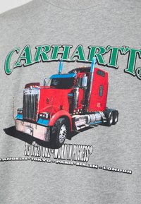 Šedé tričko s grafikou červeného nákladního vozu, s nápisem "CARHARTT" v zelené barvě nahoře a čtyřmi názvy měst níže v černé. Design zahrnuje metalické akcenty a výrazný font.
