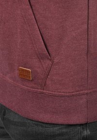 Maroon hoodie with a textured fabric, ribbed cuffs, a pocket, and a small brown leather logo patch near the hem.