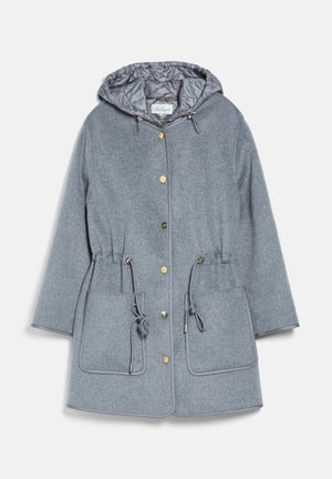 Femme portant une tenue grise en couches, comprenant une veste matelassée zippée avec capuche et un manteau en laine avec de grandes poches et des boutons dorés.