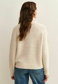 Suéter beige de punto con un corte holgado, puños y dobladillo acanalados. Textura suave, diseño ligeramente oversized, combinado con jeans azules.