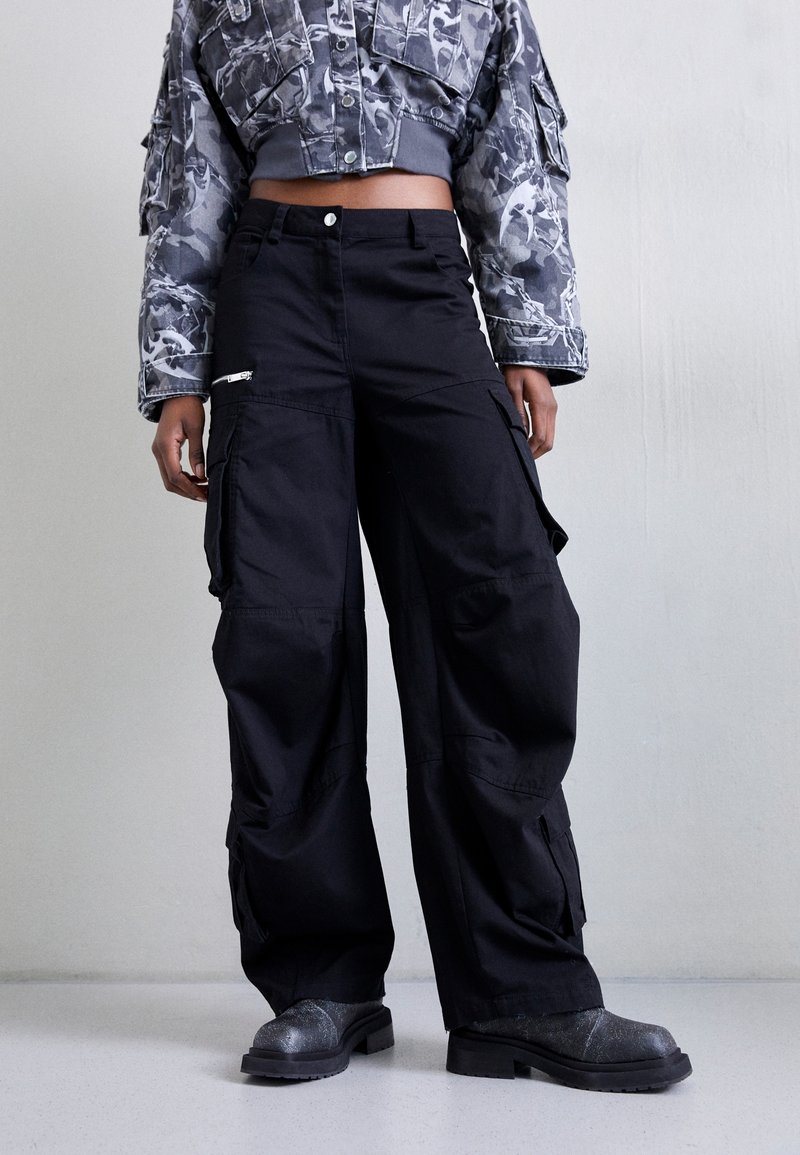 Personne portant un pantalon cargo noir à jambes larges, une veste courte camouflage et des bottes noires texturées, debout contre un mur clair uni.