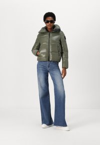Olive green puffer jacket with a high collar, zip closure, and side buttons, paired with wide-leg blue jeans and white sneakers.