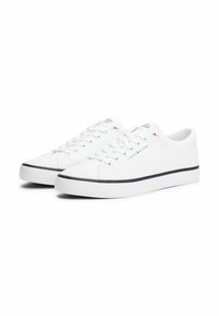 White canvas sneakers with a textured upper, round toe, flat rubber sole, and navy blue accents on the rim. Features lace-up front and logo detail.