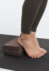 Bare feet resting on a yoga block placed on a black exercise mat, with legs wearing dark green leggings visible.