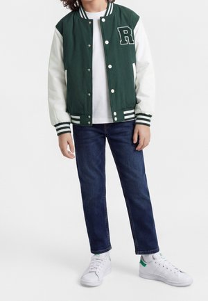 Enfant portant une veste varsity verte et blanche, un t-shirt blanc, un jean bleu foncé et des baskets blanches, debout devant un fond uni.