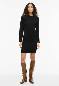 Czarna dzianinowa sukienka z długim rękawem o dopasowanym kroju. Noszona z wysokimi, błyszczącymi brązowymi skórzanymi kozakami. Neutralne tło.