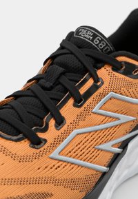 Oranje en zwarte sneaker met mesh bovenwerk, zwarte veters, reflecterende zilveren accenten en een textuurzool. Model: Fresh Foam 680.