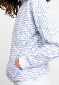 Felpa bianca con un motivo a logo ripetuto in blu "Calvin Klein", con tasca anteriore e polsini a costine. Tessuto morbido.