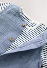 Vêtements superposés : un gilet texturé bleu avec boutons en bois sur une chemise à rayures blanches et bleu marine. Tissu doux et respirant.