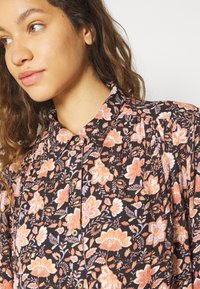 Blusa de tela negra con estampado floral. Presenta flores en tonos naranja, rosa y púrpura, con cuello de botón y detalles plisados.