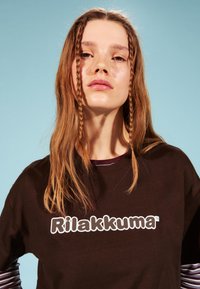 Braunes T-Shirt mit weißem Text "Rilakkuma" auf der Vorderseite. Das Model hat lange, glatte Haare mit zwei Zöpfen, die das Gesicht umrahmen, vor einem blauen Hintergrund.