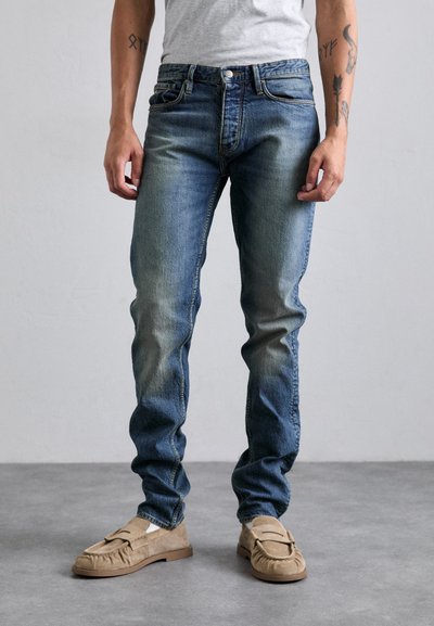 Parte inferiore del corpo di una persona che indossa jeans blu sbiaditi, mocassini in suede beige, calze bianche e una maglietta grigia, in piedi su un pavimento grigio.