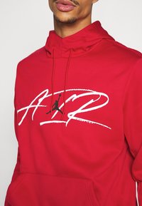 Sweat à capuche rouge avec une poche kangourou à l'avant, présentant un graphique "AIR" blanc bien visible et un logo en silhouette noire. Tissu doux et capuche réglable.
