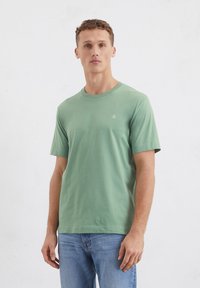 T-shirt verde in cotone con maniche corte, scollatura rotonda e un piccolo logo sul petto sinistro, indossato con jeans azzurri chiari.