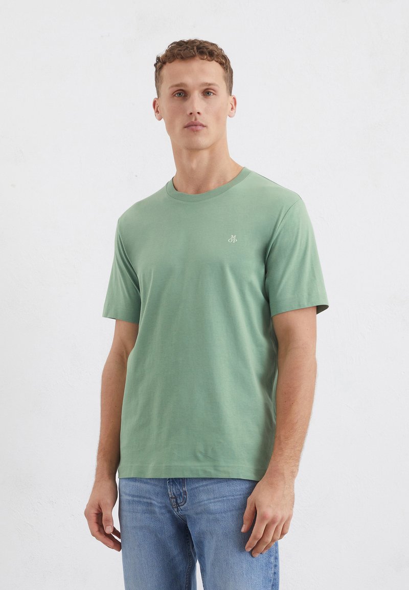 T-shirt verde in cotone con maniche corte, scollatura rotonda e un piccolo logo sul petto sinistro, indossato con jeans azzurri chiari.