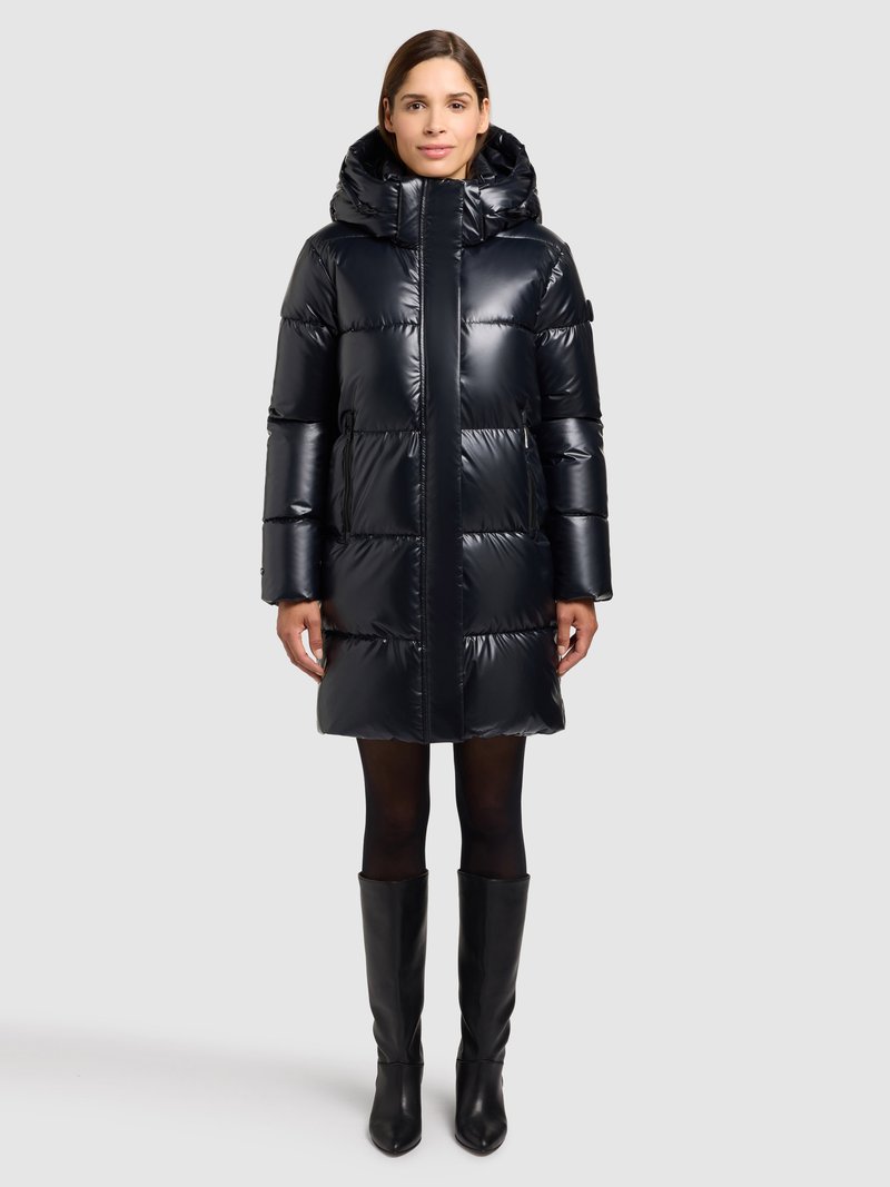 Čierny prešívaný kabát s kapucňou, s lesklým povrchom, kapucňovým dizajnom a vertikálnymi stehovými líniami. Nosený spolu s čižmami po kolená v čiernej farbe.