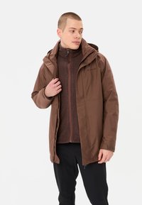 Giacca impermeabile marrone con cappuccio e tasche con zip, indossata sopra un maglione marrone strutturato, abbinata a pantaloni neri.