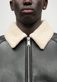 Czarna skórzana kurtka z kołnierzem z kremowego sztucznego futra, z metalowym zamkiem błyskawicznym i gładką fakturą. Zbliżenie, skupione na detalach.