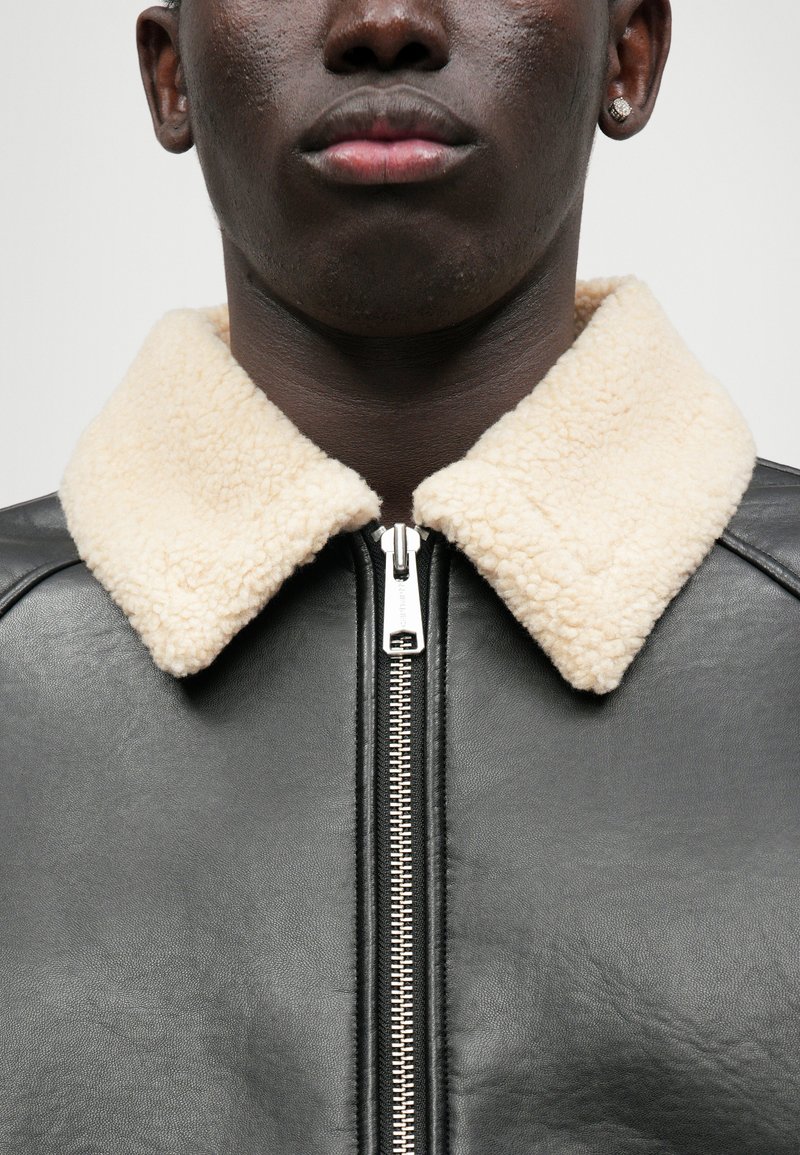 Schwarze Lederjacke mit einem Kragen aus cremefarbenem Kunstfell, ausgestattet mit einem silbernen Reißverschluss und glatter Textur. Nahaufnahme, fokussiert auf Details.