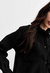 Camicia nera oversize realizzata in tessuto morbido, con chiusura a bottoni sul davanti, due tasche sul petto e maniche voluminose.