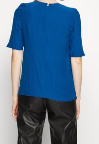 Niebieska bluzka z krótkim rękawem, okrągłym dekoltem, lekko rozkloszowanymi rękawami i zamkiem błyskawicznym z tyłu. Miękki materiał o gładkiej fakturze.