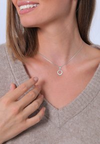 Collier en argent avec un pendentif circulaire, ornée d'une pierre claire, porté avec un pull beige à col en V, les mains touchant délicatement le collier.