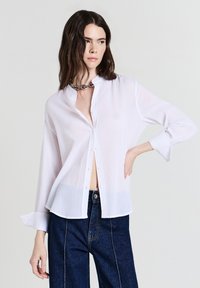 Camicia bianca con colletto alla coreana, maniche lunghe e un tessuto leggero e texturizzato. Presenta una vestibilità leggermente rilassata e un orlo arrotondato.