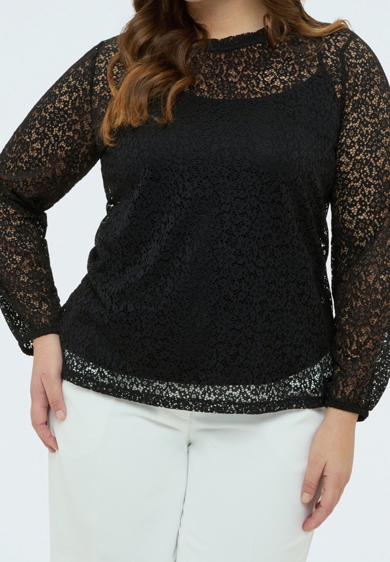 Femme portant un haut en dentelle noire à manches longues par-dessus un caraco noir, associé à un pantalon blanc, sur un fond clair uni.