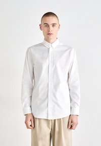 Chemise blanche à manches longues avec un col pointu, fermeture à boutons et texture lisse, assortie à un pantalon beige. Design simple et épuré.