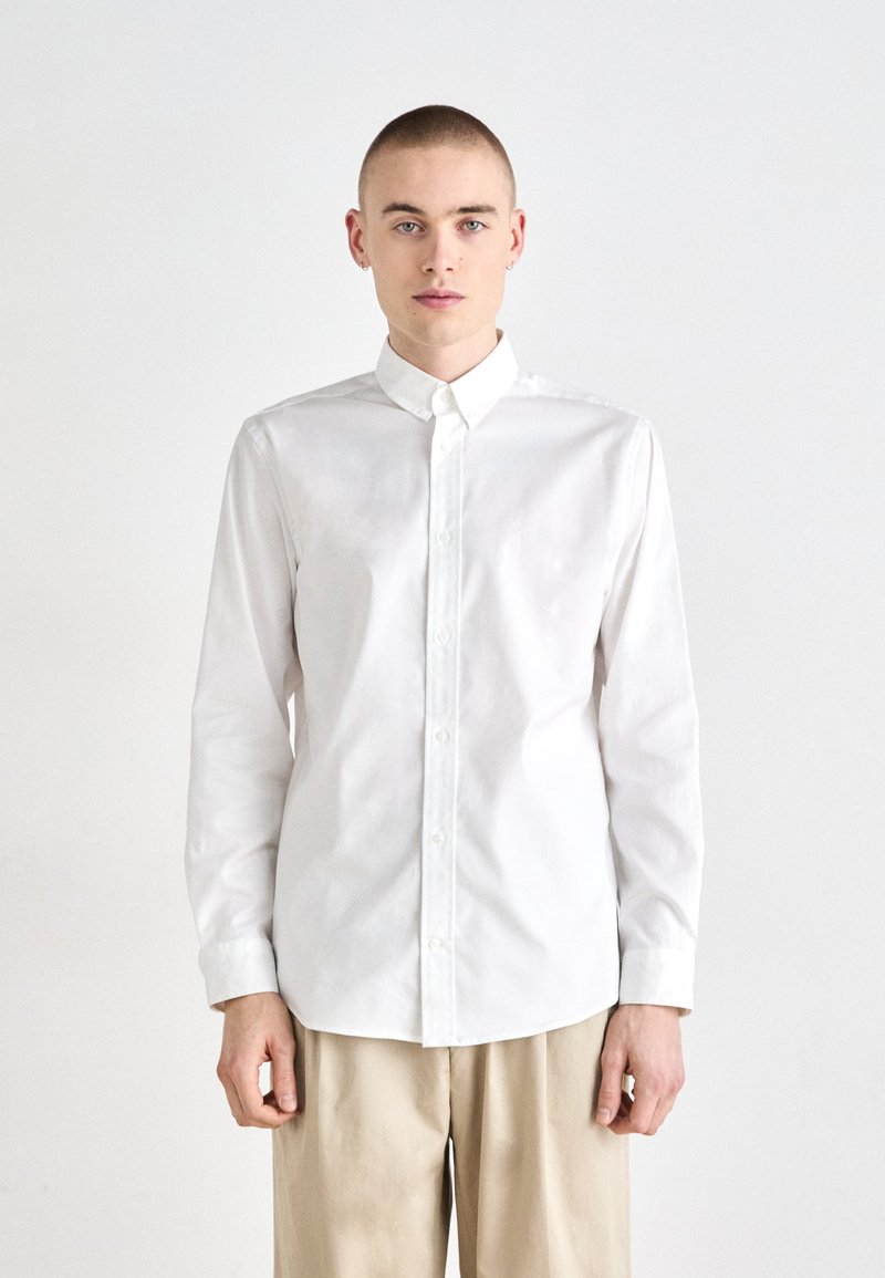 Chemise blanche à manches longues avec un col pointu, fermeture à boutons et texture lisse, assortie à un pantalon beige. Design simple et épuré.