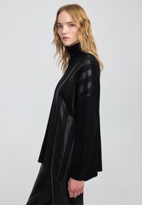 Młoda kobieta z falującymi blond włosami nosi czarny sweter z golfem z błyszczącymi pionowymi paskami i czarne skórzane spodnie, stojąc bokiem.