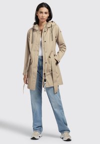 Beige, waterproof jacket with a hood, drawstrings, snap buttons, and side pockets. Paired with light blue jeans and neutral sneakers.