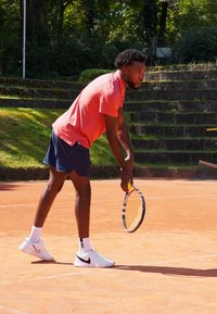 Male tennis player in a pink shirt and navy shorts, preparing to serve on a clay court, holding a racket, wearing white athletic shoes.