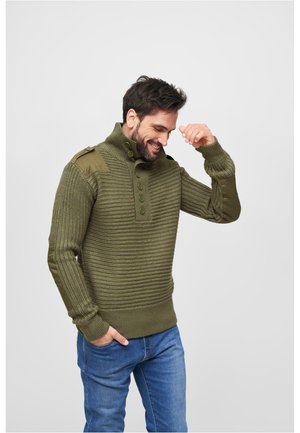 Pulover tricotat de culoare verde măslin, cu guler înalt, deschidere butonată, manșete cu bordură și panouri contrastante pe umeri. Purtați cu blugi albaștri.
