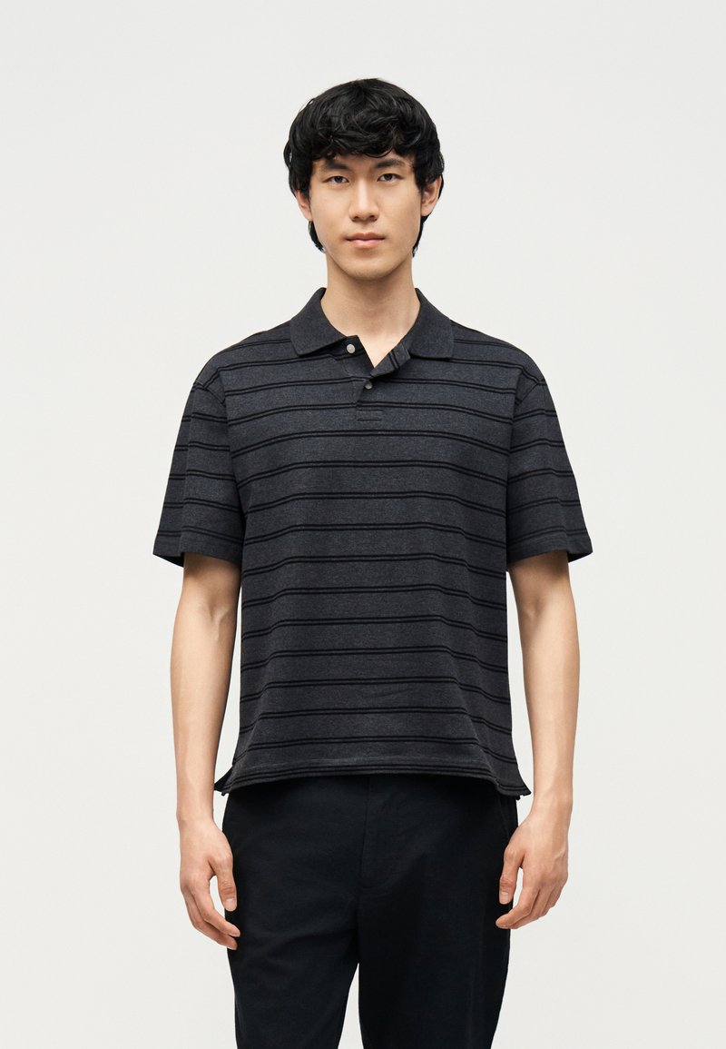 Νέος άνδρας που φοράει κοντομάνικη πόλο μπλούζα με οριζόντιες μαύρες και σκουρόγκρι ρίγες και μαύρο παντελόνι, στέκεται μπροστά σε απλό ανοιχτό φόντο.