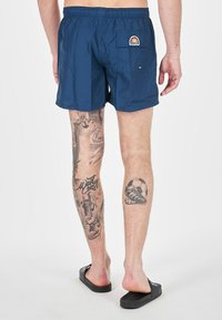 Bañadores azul marino con cinturilla elástica y un bolsillo trasero, con un colorido parche de la marca. Llevados con sandalias oscuras; tatuajes notables en las piernas.