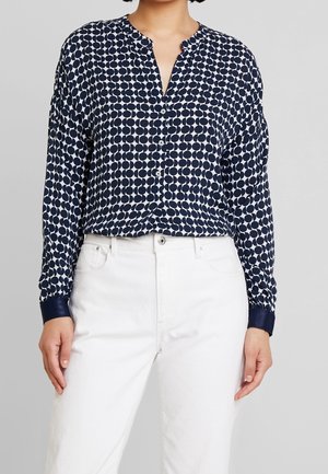 Kobieta ubrana w granatowo-białą koszulę zapinaną na guziki z wzorem w kółka, z granatowymi mankietami oraz białe spodnie z wysokim stanem, na jednolitym tle.