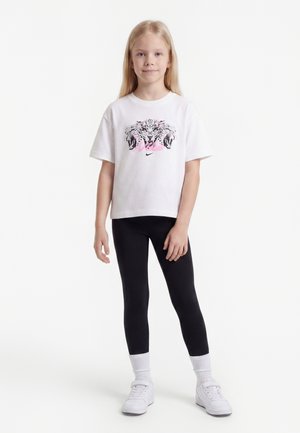 Blond pige står, iført en hvid t-shirt med leopardmotiv, sorte leggings, hvide sokker og hvide sneakers på en hvid baggrund.