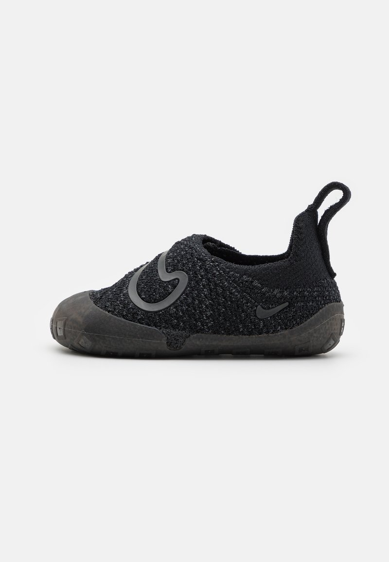 Svart textil sko med slip-on-design, strukturerad ovandel, gummisula och framträdande logotypaccent på sidan. Har en dragflik på hälen.