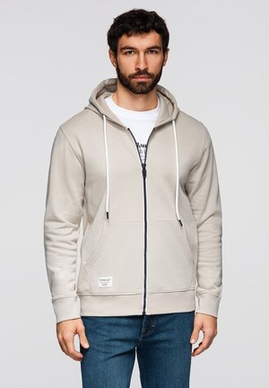 Homme aux cheveux foncés et à la barbe, portant un sweat à capuche beige zippé sur un t-shirt blanc et un jean bleu, debout devant un fond uni.
