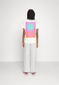 Personne avec des cheveux tressés portant un pantalon blanc, des chaussures lilas, et un t-shirt blanc avec un graphic coloré de soleil et le texte "ESSENCE D'ÉTÉ" au dos.