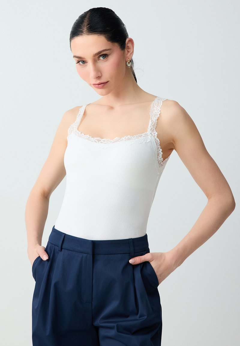 Femme aux cheveux foncés et boucles d'oreilles créoles dorées portant un débardeur blanc bordé de dentelle et un pantalon taille haute bleu marine, les mains dans les poches.
